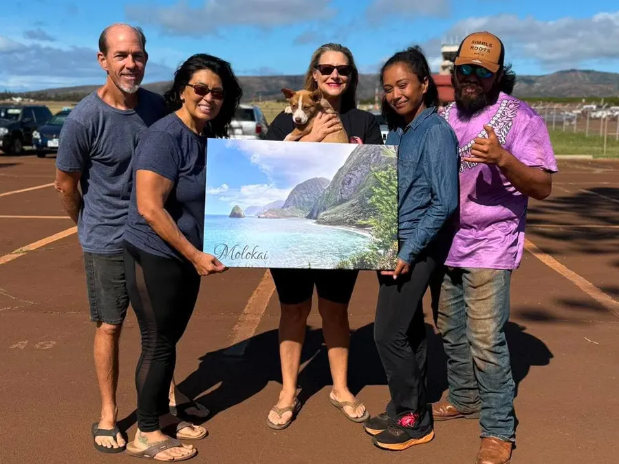 Five people at the Molokaʻi airport, one holding a puppy, with a picture of the Molokaʻi coast